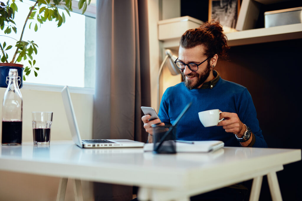 bearded entrepreneur sitting in his office, using smart phone and taking a break from work.