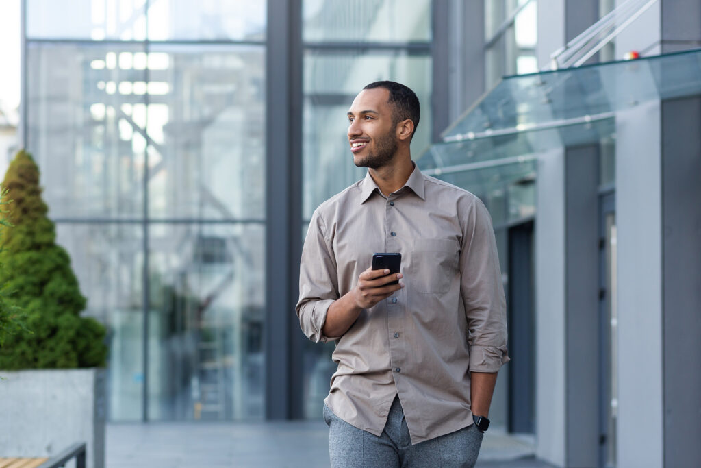 An adult man in a beige shirt and gray trousers, walking near a contemporary glass office building while looking at his smartphone with a smile.