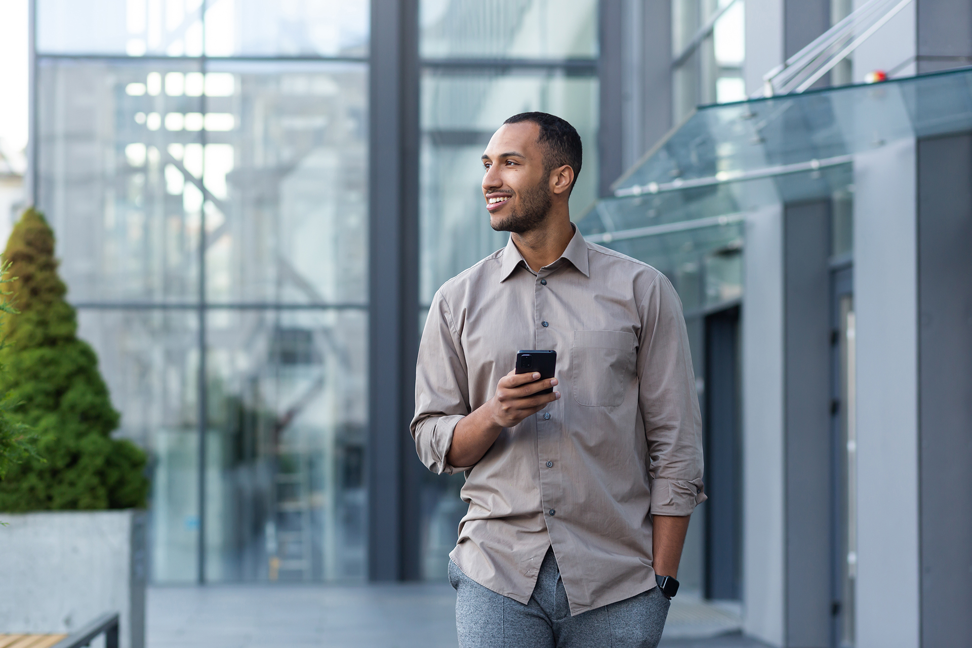 An adult man in a beige shirt and gray trousers, walking near a contemporary glass office building while looking at his smartphone with a smile.