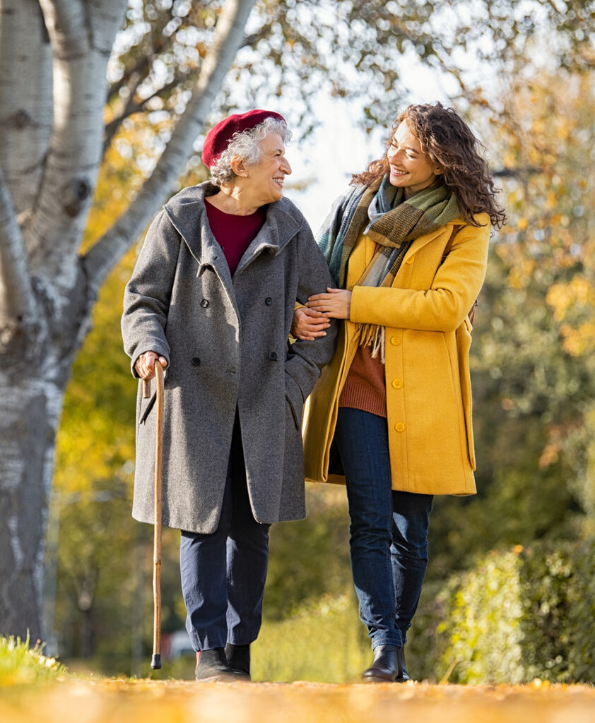 Granddaughter walking with senior woman in park wearing winter clothing. Old grandmother with walking cane walking with lovely caregiver girl in sunny day. Happy woman and smiling grandma walking in autumn park.