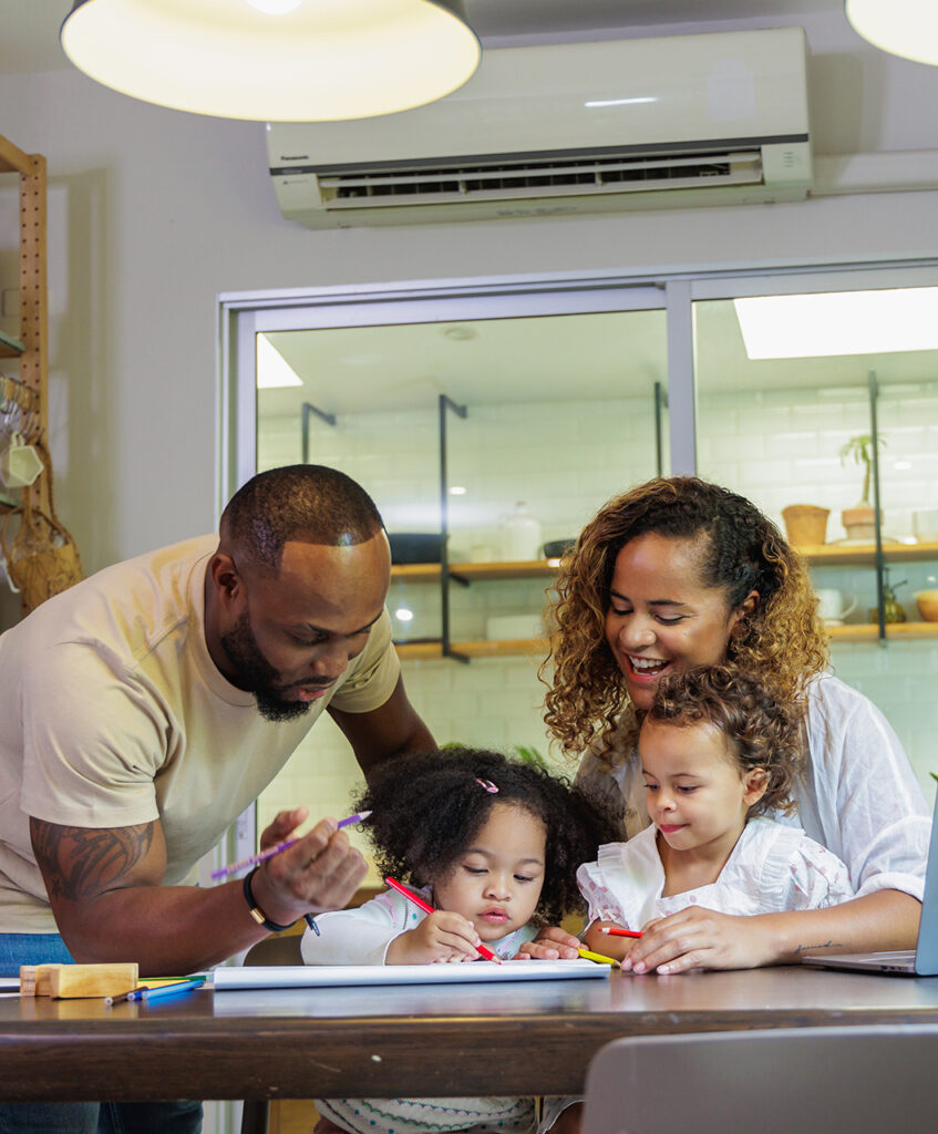 Two adults and two young children sharing smiles while drawing together at a table in a warmly lit kitchen or dining area.