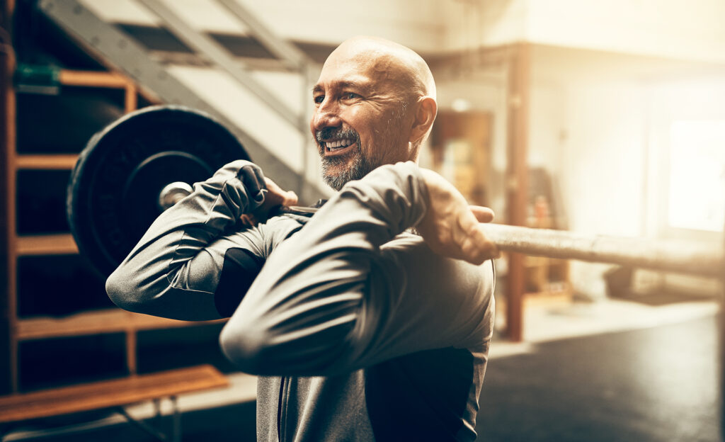 Fit mature man smiling while standing in a gym lifting heavy weights during a workout session