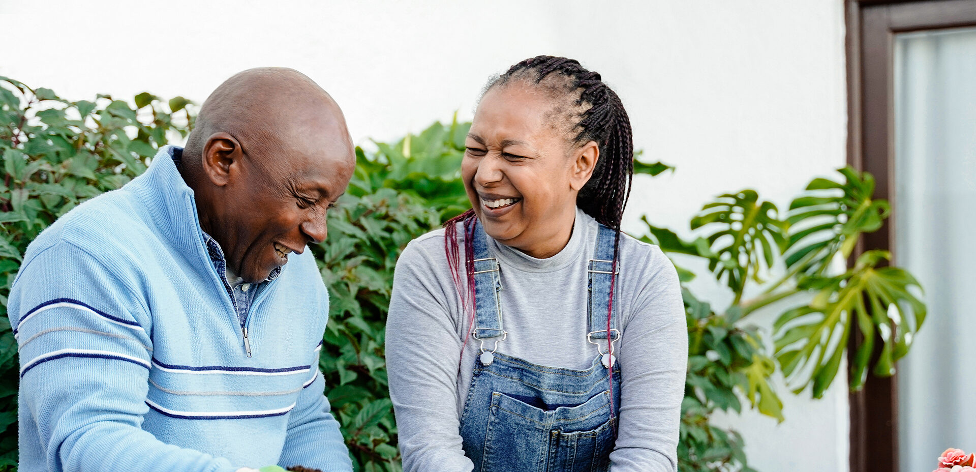Older black couple laughing together while repotting flowers and herbs on a patio.